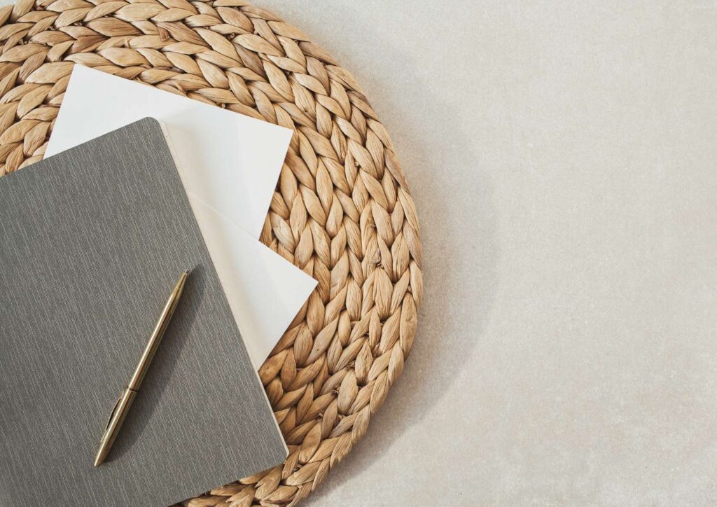 Notebook and gold pen resting on a woven placemat, symbolising reflection, clarity, and rewriting your personal money story.