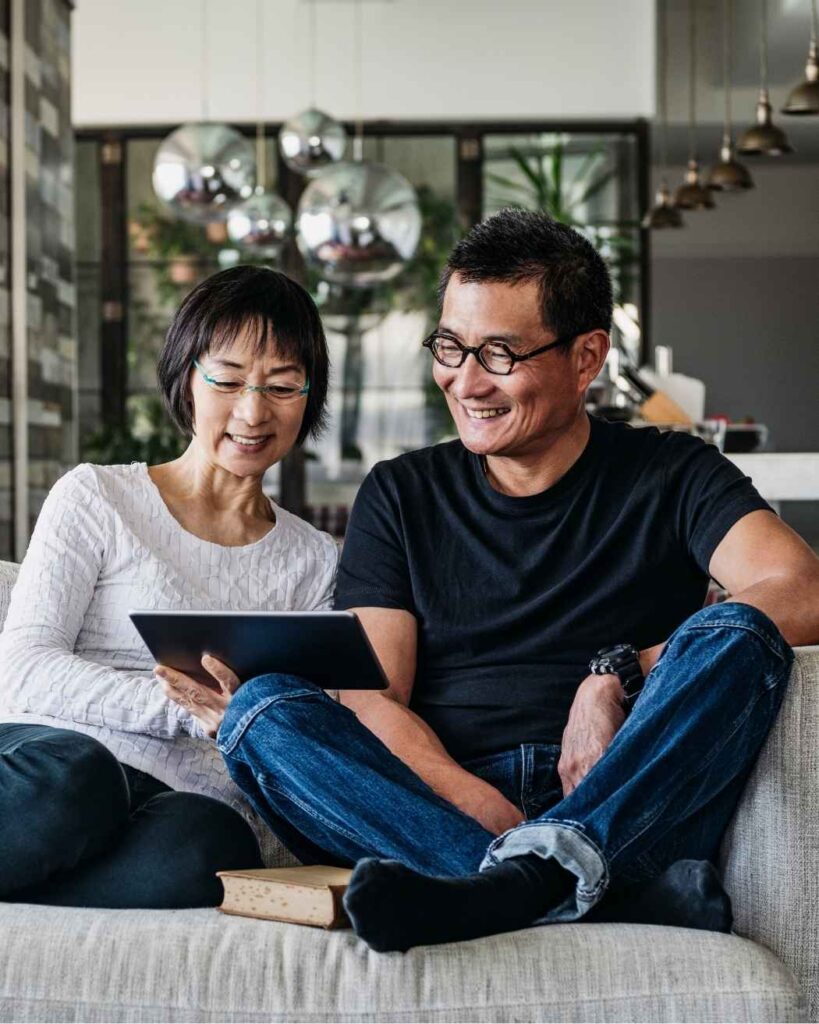 Smiling mature couple sitting on sofa looking at a tablet, representing confident retirement planning and financial peace of mind.