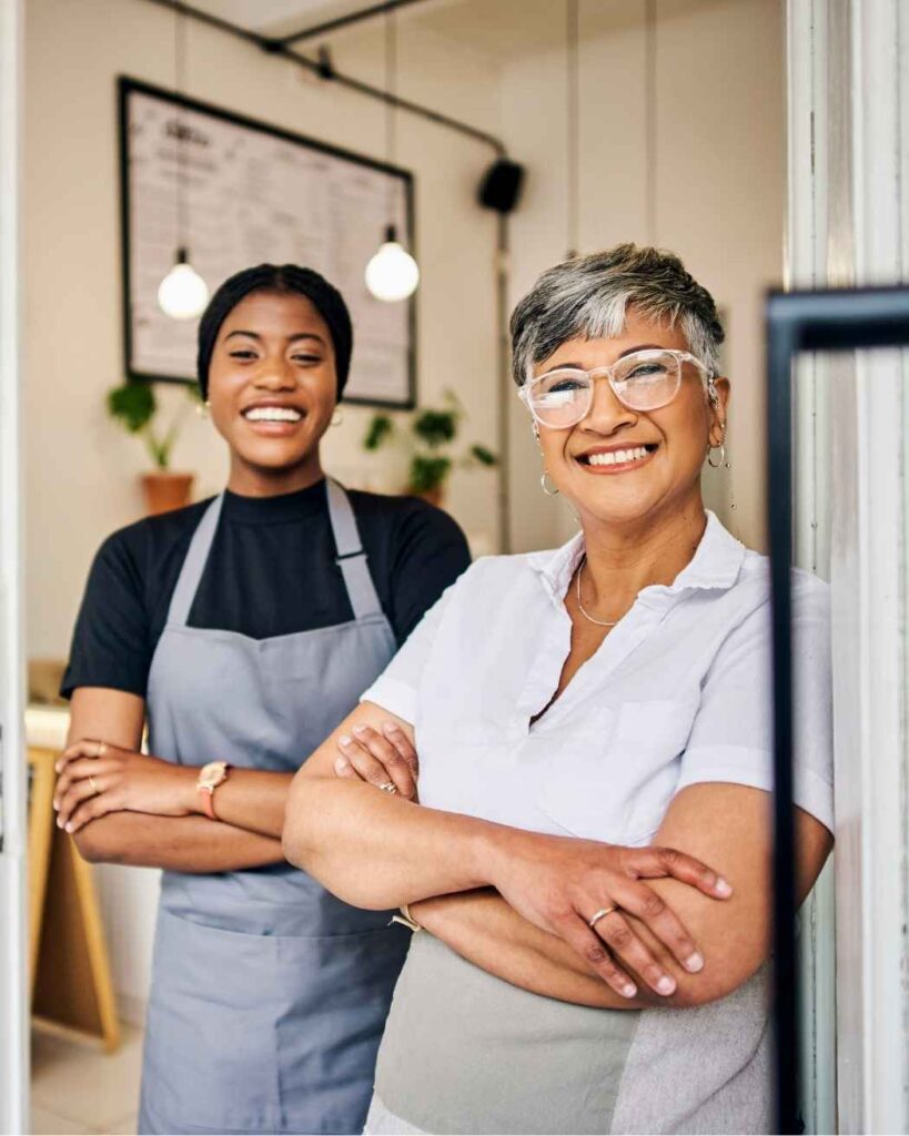 Two business women in aprons smiling in a cafe, representing real Ikigai Wealth clients living with financial clarity and confidence.